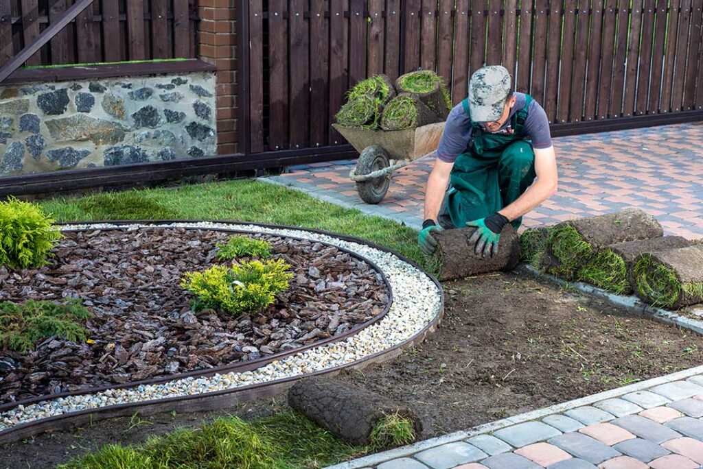 A person assembling a wooden raised garden bed frame with soil and tools nearby in a green backyard.