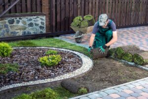 A person assembling a wooden raised garden bed frame with soil and tools nearby in a green backyard.