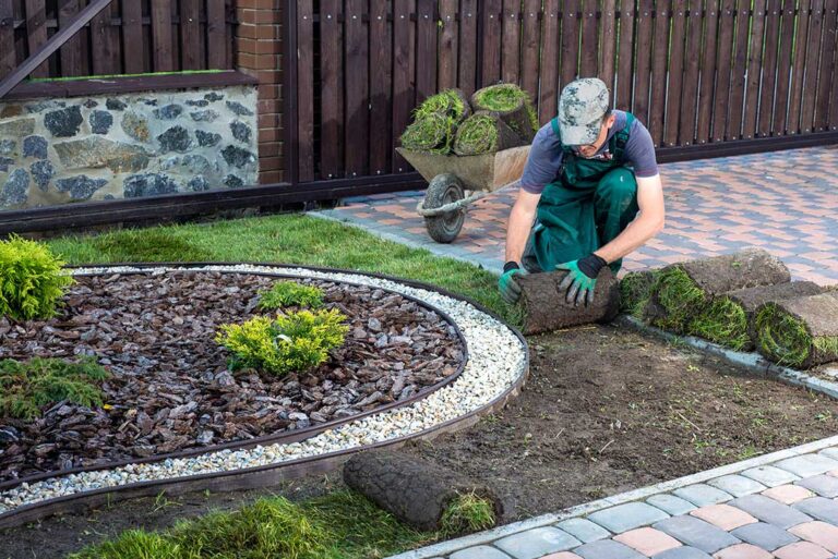 A person assembling a wooden raised garden bed frame with soil and tools nearby in a green backyard.